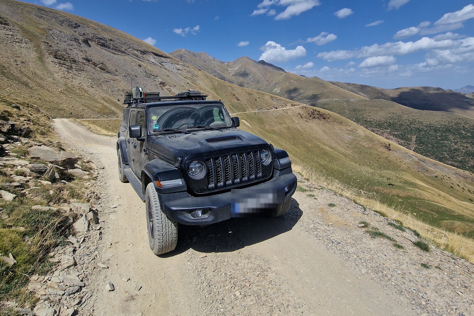Shadow, the black Jeep, on a gravel track in the Pyrenees with mountain ridges and blue sky behind