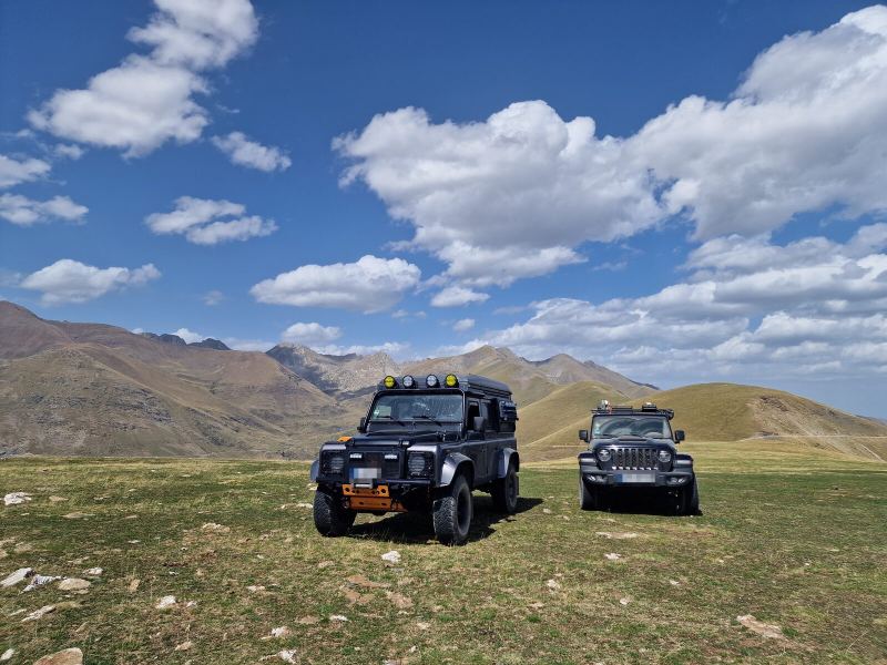 Der Defender D110 und Shadow nebeneinander auf einer Hochweide, Wolken über den Gipfeln